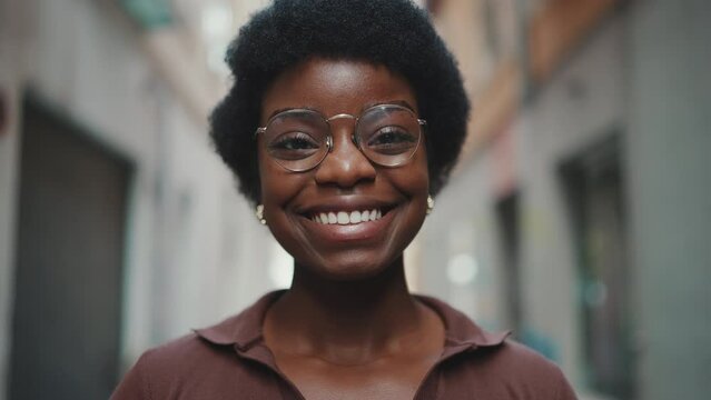 Close up shot of beautiful African woman in glasses looking happy outdoors. Pretty dark-skinned girl looking at camera sincerely smiling on the city street. Face expression concept