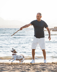 60-years old man playing with small cute jumping and barking dog jack russell terrier on sea beach