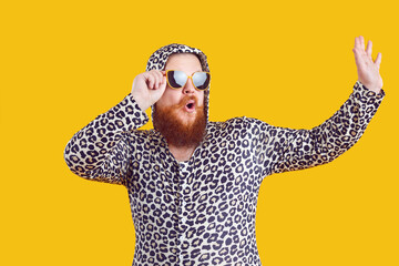Portrait of funny comic chubby man who is strangely dressed and having fun on orange background. Cheerful crazy millennial bearded fat man dressed in leopard sweater with hood and sunglasses.
