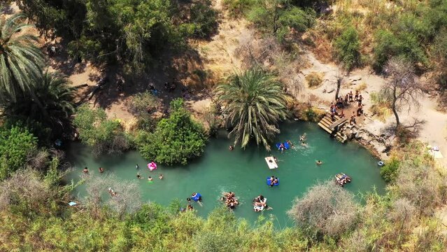 Many people float on air mattresses on a warm sun-drenched day on the clear blue natural spring while young people enthusiastically splash from a platform into the water. High angle drone panning shot