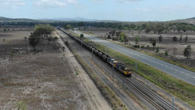 Empty coal train running on railway parallel to road, Bajool in Queensland, Australia. Aerial backward