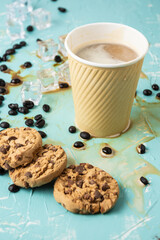 Top view of a cardboard cup with cappuccino coffee on a blue table, spilled coffee, coffee beans and cookies , vertical, with copy space