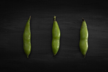 three broad beans in shell lined up on a black wooden background