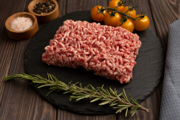 Minced meat on a cutting board with a sprig of rosemary, cherry tomatoes, pink salt and a mixture of peppers on a wooden background, flat lay, open space
