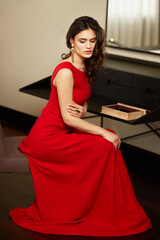 Fototapeta premium Gorgeous young woman with brown wavy hair in elegant dress sitting on chair in luxury interior