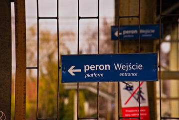 Tabliczka informacyjna ( strzałka kierunkowa ) o treści : " peron wejście  platform entrace " . Biały napis na niebieskim tle .Tabliczka wisząca na podrdzewiałym płocie ze stalowych prętów na dworcu . © Grzegorz