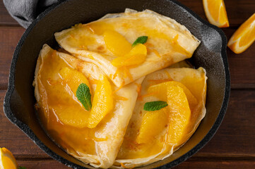 Delicious crepes suzette with orange syrup and slices fruits on a frying pan on a dark wooden background, top view
