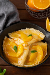 Delicious crepes suzette with orange syrup and slices fruits on a frying pan on a dark wooden background, top view
