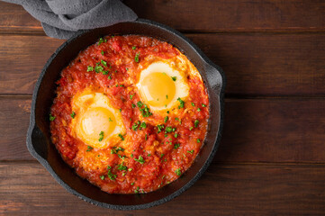 Shakshuka in a frying pan on a dark wooden background. Eggs cooked in tomato sauce with spices and fresh parsley on top. Rustic style. Top view, copy space.