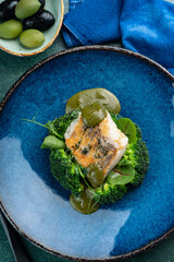 Fried cod fillet with broccoli, herbs and basil sauce on a blue plate, top view