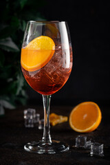 Shining cocktail Aperol syringe in a wine glass with ice and a slice of orange on a dark background with tree leaves