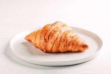 Plate with one fresh croissant on white wooden table.