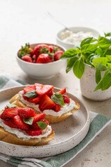 Homemade bruschettas with strawberries, cream cheese and basil in white plate on light background. Summer appetizer. Selective focus.