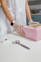 female hands in gloves ready to decorate pink box of cupcakes with white ribbon, scissors on white background. Valentines day present