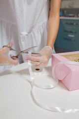 female hands in gloves cutting with scissors white ribbon to decorate pink box of cupcakes on white table background. Mothers day present