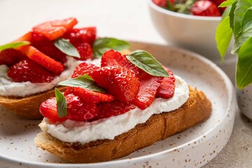 Homemade bruschettas with strawberries, cream cheese and basil in white plate on light background. Summer appetizer. Selective focus.