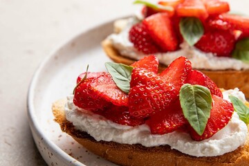 Homemade bruschettas with strawberries, cream cheese and basil in white plate on light background. Summer appetizer. Selective focus.