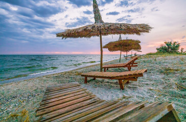 Beautiful sunrise or sunset over the calm waters of the Black Sea on the Kinburg Spit in Ukraine. Beach summer landscape. Straw umbrella and wooden deck chair on the beach at sunset or sunrise