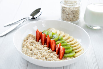Oatmeal porridge with fresh fruits, strawberry, banana, kiwi in white bowl with glass of milk, jar with oats and spoon on background. Recipe of healthy breakfast, clean eating.