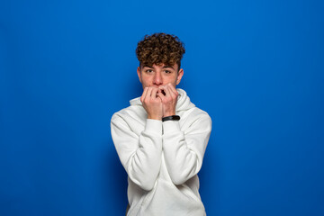 Fototapeta na wymiar Young caucasian man with curly hair isolated for blue background dressed in a white sweatshirt, covering his mouth with his hands and round eyes experiencing deep amazement and fear.