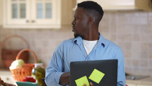 Thoughtful concentrated African American man surfing Internet on laptop searching recipe online. Portrait of focused young guy thinking looking back at fruits and vegetables