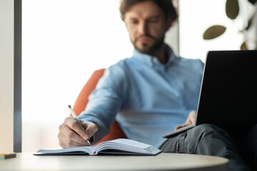 Close up of man using laptop and writing in notebook