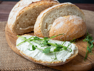 Close-up of a still life made of fresh crusty bread. A slice of bread smeared with cottage cheese and decorated with micro greenery. Linen napkin brown wooden background, top view, flat lay