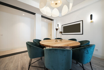 Round dining table in raw wood varnish surrounded by blue velvet upholstered armchairs under a designer lamp