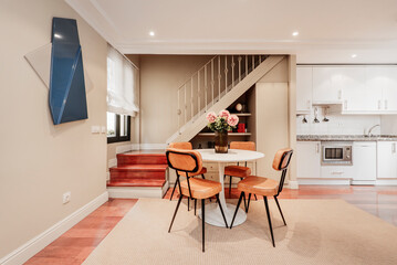 Living room with round white dining table with tobacco colored leatherette upholstered chairs, open plan kitchen with white cabinets and staircase to attic bedrooms