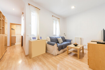 Living room with a folded wooden dining table, a two-seater sofa, various wooden furniture, a TV on a chest of drawers and a corridor leading to a kitchen with shelves and curtained windows