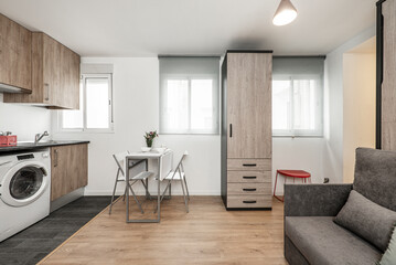 Studio apartment with an open kitchen, a living room with a gray fabric upholstered folding sofa bed, a wardrobe with drawers and a white folding dining table with matching chairs and a wooden floor