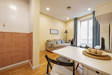 Studio apartment with white folding dining table with matching black chairs, three-seater sofa bed upholstered in beige fabric and balcony with glass and metal and blue curtains