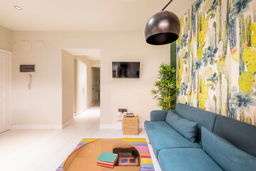 Living room with a three-seater sofa in blue fabric, a wall with decorative wallpaper in bright colors and an oblong glass coffee table