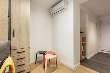 Corner of a living room with colored side chairs, air conditioner, wooden cabinet with drawers and multi-colored flooring