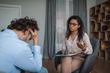 Serious young african american lady psychologist consulting crying depressed european man in clinic