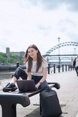 Fototapeta premium A cheerful girl works with a laptop on the embankment sitting on a bench. The concept of an open-air office. Freelancer at work. Newcastle, Time Bridge, North East England.