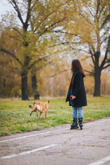 A young girl in jeans, a coat and a long scarf is skating in an autumn park, along which a street dog is running. Fighting autumn depression.