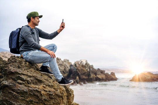 latin man taking pictures with smartphone and smiling on beach happy