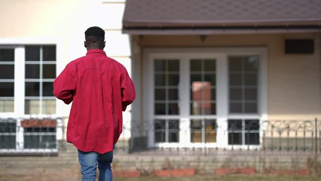 Back view young African American man walking to house outdoors on spring summer day. Confident millennial owner strolling on front yard moving in new home