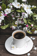 A beautiful postcard. A white coffee cup with a saucer, a statuette, candles, a book and a vase with a bouquet of blooming apple trees. Beautiful still life. Spring time. The concept of 