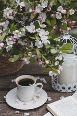 A beautiful postcard. A white coffee cup with a saucer, a statuette, candles, a book and a vase with a bouquet of blooming apple trees. Beautiful still life. Spring time. The concept of 