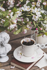 A beautiful postcard. A white coffee cup with a saucer, a statuette, candles, a book and a vase with a bouquet of blooming apple trees. Beautiful still life. Spring time. The concept of 