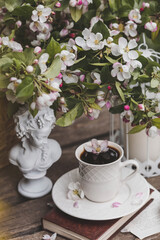 A beautiful postcard. A white coffee cup with a saucer, a statuette, candles, a book and a vase with a bouquet of blooming apple trees. Beautiful still life. Spring time. The concept of 