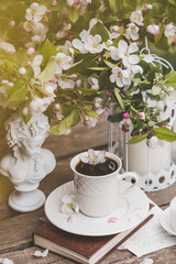 A beautiful postcard. A white coffee cup with a saucer, a statuette, candles, a book and a vase with a bouquet of blooming apple trees. Beautiful still life. Spring time. The concept of 