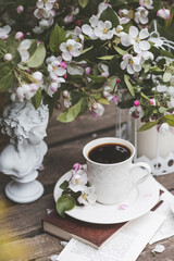 A beautiful postcard. A white coffee cup with a saucer, a statuette, candles, a book and a vase with a bouquet of blooming apple trees. Beautiful still life. Spring time. The concept of 