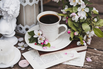 A beautiful postcard. A white coffee cup with a saucer, a statuette, candles, a book and a vase with a bouquet of blooming apple trees. Beautiful still life. Spring time. The concept of 