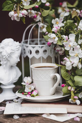 A beautiful postcard. A white coffee cup with a saucer, a statuette, candles, a book and a vase with a bouquet of blooming apple trees. Beautiful still life. Spring time. The concept of 