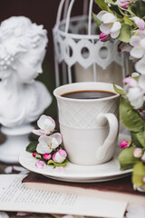 A beautiful postcard. A white coffee cup with a saucer, a statuette, candles, a book and a vase with a bouquet of blooming apple trees. Beautiful still life. Spring time. The concept of 