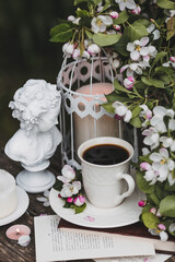 A beautiful postcard. A white coffee cup with a saucer, a statuette, candles, a book and a vase with a bouquet of blooming apple trees. Beautiful still life. Spring time. The concept of 