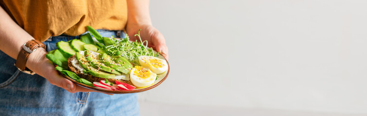 A woman holds a plate with useful healthy foods avocado, cucumber, spinach, radish, egg, microgreens